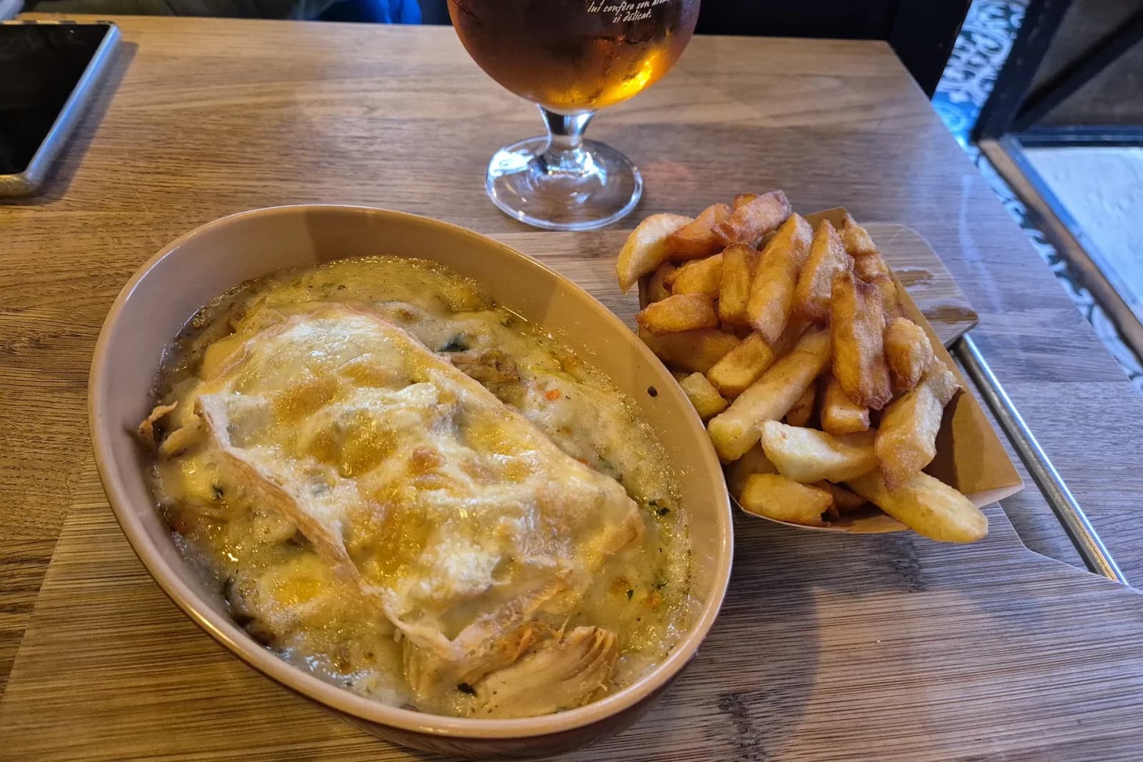 Un gratin de poulet au maroilles dans son plat à oreilles, avec un cornet de frites et une bière ambrée.