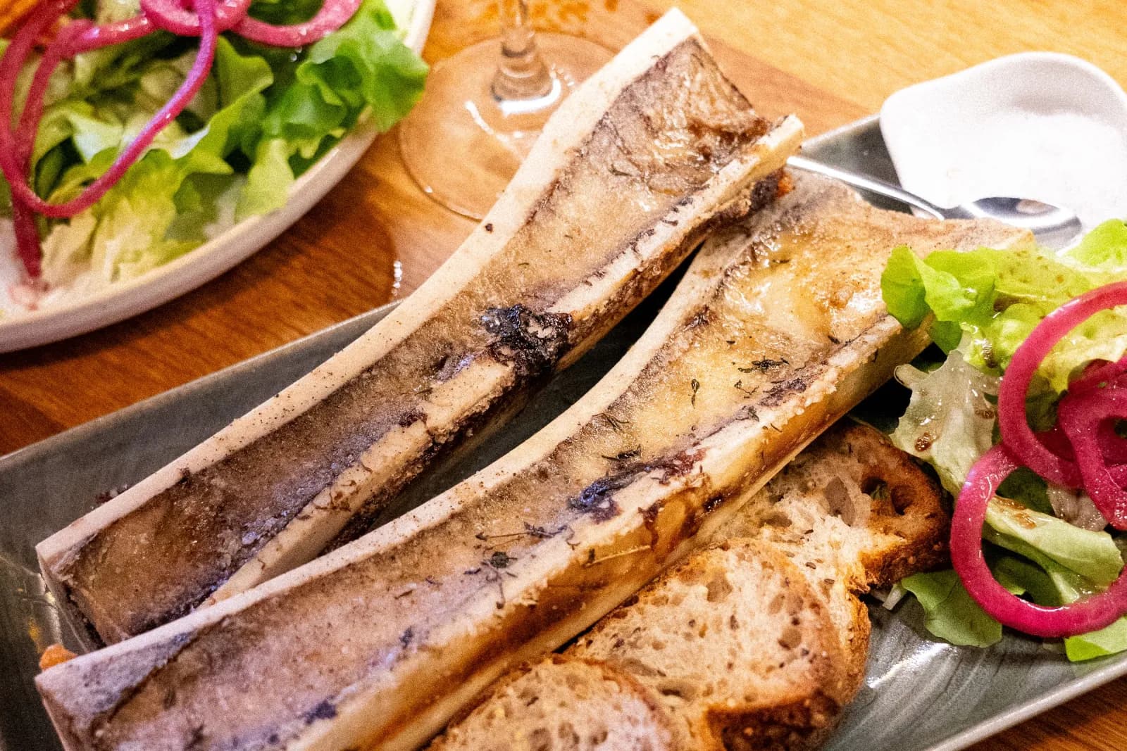 Des os à moelle servis sur une ardoise avec une salade et une bière, sur une table de bois.